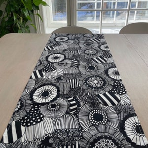 Könnte beinhalten: Ein Tischläufer mit schwarz-weißem Blumenmuster. Der Läufer zeigt ein sich wiederholendes Design aus stilisierten Blumen und geometrischen Formen. Der Läufer liegt auf einem hellen Holztisch und erzeugt einen auffälligen Kontrast. Das Design ist kühn und grafisch.