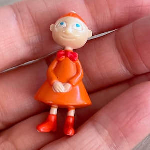 May include: A small, orange figurine of a cartoon character with a red bow tie, red boots, and a matching cap. The figurine has a smiling face with blue eyes and is holding its hands together. The figure is approximately 2 inches tall.
