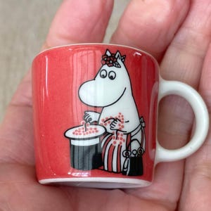 Può includere: Una tazza in ceramica rossa in miniatura con un personaggio Moomin bianco che indossa una corona di fiori. Il personaggio è raffigurato accanto a un cilindro nero con puntini rossi, mentre tiene in mano un piccolo oggetto rosso. Interno e manico bianchi.