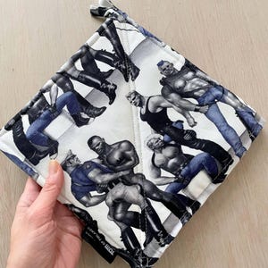 May include: A quilted pot holder with a black and white print featuring illustrations of muscular figures in various poses. The pot holder has a white background and a black border. The tag reads "TOTTI OF FINLAND".