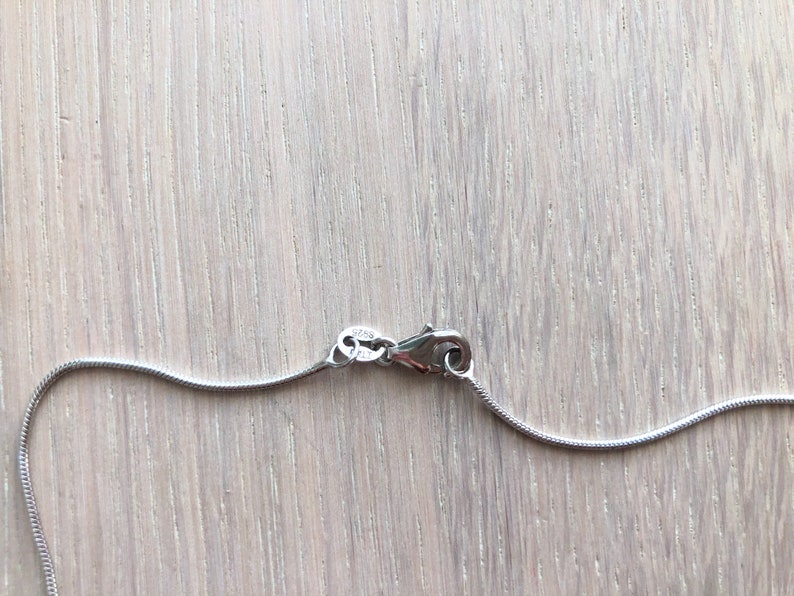 Pode incluir: Um close-up de um colar de prata com um design de corrente de cobra. O colar &eacute; preso com um fecho lagosta. O fecho est&aacute; conectado &agrave; corrente. O fundo &eacute; uma superf&iacute;cie de madeira clara.