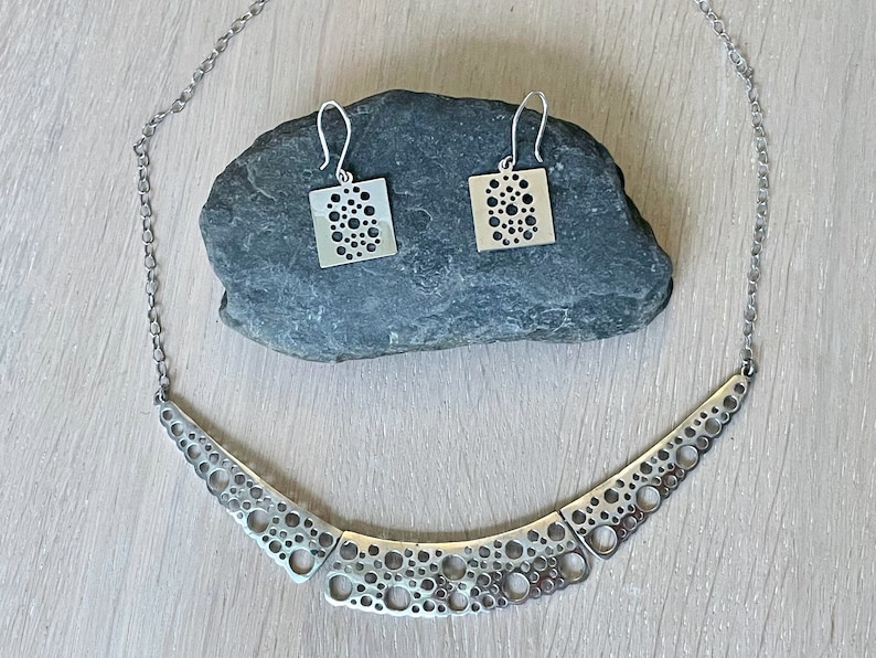 May include: A silver necklace and earrings set with a geometric design featuring a pattern of small circles. The necklace is a curved shape with multiple layers of circles.