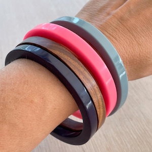 Puede incluir: Un conjunto de cuatro pulseras apiladas en una muñeca. Las pulseras están hechas de diferentes materiales, incluyendo madera, plástico y resina. Los colores de las pulseras son rosa, azul, marrón y negro.