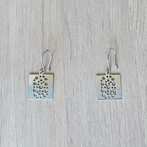 May include: A pair of silver earrings with a square shape and a cutout design of small circles.