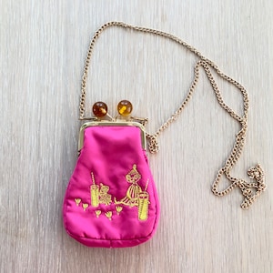 May include: A bright pink satin purse with a gold chain strap. The purse has a gold clasp and amber-colored ball closures. The front of the purse features gold embroidered figures and designs.