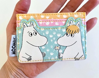 May include: A colorful card holder featuring a Moomin design. The card holder has a light blue background with Moomin characters, a yellow, pink, and light blue top, and the word "MOOMIN" on a white tag.
