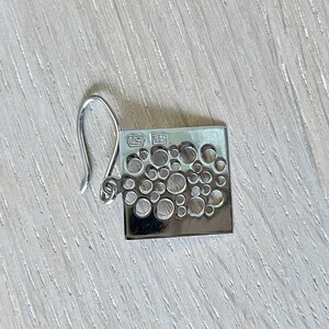 May include: Silver earring with a square shape and a design of small circles cut out. The earring has a hook fastening and is marked "925" on the back.