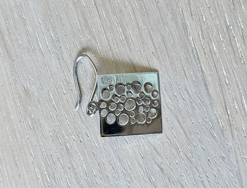 May include: Silver earring with a square shape and a design of small circles cut out. The earring has a hook fastening and is marked "925" on the back.