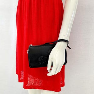 Pode incluir: Uma carteira de m&atilde;o de couro preto com um fecho de correr prateado e uma pequena al&ccedil;a de corrente prateada. A carteira tem uma superf&iacute;cie texturizada e um design em relevo subtil.