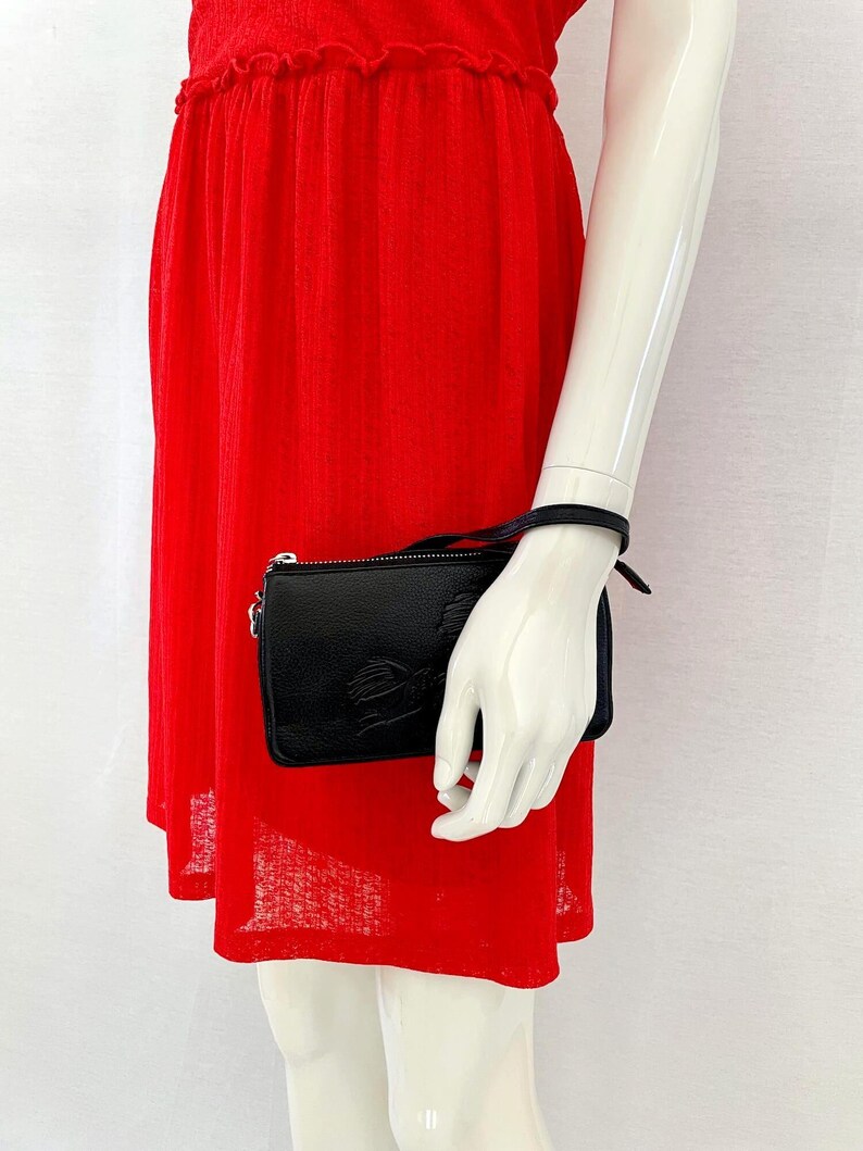 Pode incluir: Uma carteira de m&atilde;o de couro preto com um fecho de correr prateado e uma pequena al&ccedil;a de corrente prateada. A carteira tem uma superf&iacute;cie texturizada e um design em relevo subtil.