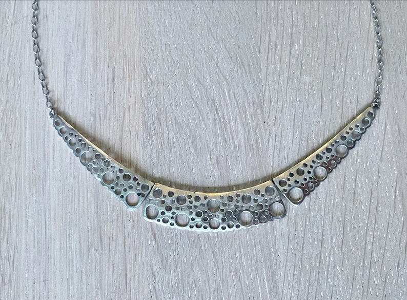 May include: A silver necklace with a geometric design featuring a series of interconnected circles. The necklace is made of a metal chain and has a modern, minimalist style.