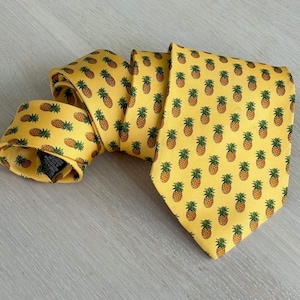 Può includere: Cravatta gialla con un motivo ripetuto di ananas marroni e verdi. La cravatta è realizzata in un tessuto liscio e si trova su una superficie grigio chiaro.