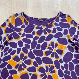 May include: A long-sleeved purple top with a floral pattern in orange and light blue. The top has a scoop neckline and a relaxed fit. The fabric appears to be a soft, lightweight material. The overall design is modern and eye-catching.