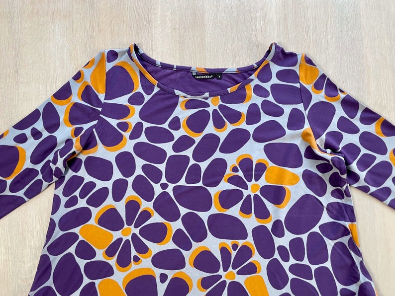 May include: A long-sleeved purple top with a floral pattern in orange and light blue. The top has a scoop neckline and a relaxed fit. The fabric appears to be a soft, lightweight material. The overall design is modern and eye-catching.