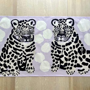 Peut inclure: Un chemin de table rectangulaire en tissu sur fond lilas clair, orné de deux léopards illustrés. Chaque léopard est blanc avec des taches noires et des traits du visage détaillés. Le motif inclut des formes blanches rappelant des nuages.