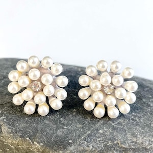 Op de afbeelding: Een paar parelcluster-oorbellen. Elke oorbel heeft een centrale heldere strass-steen omringd door talrijke kleine, ronde, crèmekleurige parels, die een bloemen- of sterpatroon vormen. De oorbellen zijn gezet op een lichtroze basis.