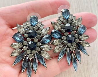 May include: A pair of statement earrings featuring a floral design. The earrings showcase a combination of blue, gray, and clear crystals. The crystals are arranged in a symmetrical pattern, with a central cluster and radiating petals.