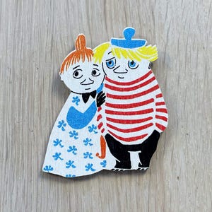 May include: A wooden figurine set of two characters from the Moomin series. The female character is wearing a blue and white dress with a red bow. The male character is wearing a red and white striped shirt with a blue hat.