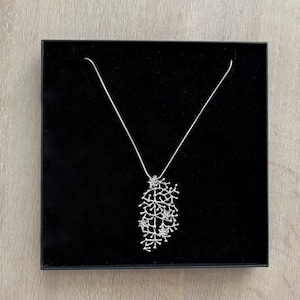 Pode incluir: Um colar prateado com um pendente ramificado e detalhado, apresentado numa caixa de joias preta. O pendente apresenta um design org&acirc;nico complexo, semelhante a um grupo de pequenos ramos ou coral. O colar &eacute; exibido numa superf&iacute;cie de madeira clara.