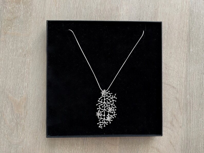 Pode incluir: Um colar prateado com um pendente ramificado e detalhado, apresentado numa caixa de joias preta. O pendente apresenta um design org&acirc;nico complexo, semelhante a um grupo de pequenos ramos ou coral. O colar &eacute; exibido numa superf&iacute;cie de madeira clara.