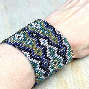 May include: A wide beaded cuff bracelet in shades of blue, green, gold, and white. The bracelet features a geometric pattern and is worn on a wrist. The beads are small and tightly woven together.