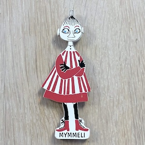 May include: A wooden figurine of a character from the Moomin stories. The character is wearing a red and white striped dress and black shoes. The figurine is standing on a wooden surface and has the name "Mymmeli" written on the base.