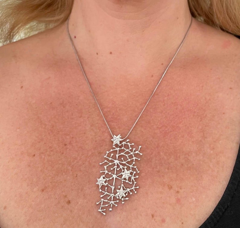 Pode incluir: Um colar prateado com um pendente abstrato. O pendente apresenta uma rede de linhas interligadas com detalhes em forma de estrela. O colar &eacute; suspenso por uma fina corrente prateada. O design &eacute; elegante e moderno.