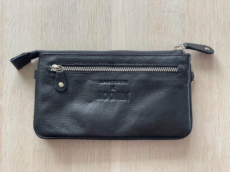 Pode incluir: Carteira de couro preta com fecho de correr e o texto "Moschino" em relevo na frente.