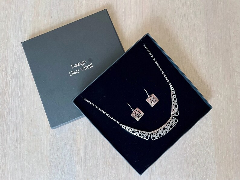 May include: A silver necklace and earrings set in a black box. The necklace has a geometric design with a pattern of circles. The earrings are square shaped with the same circle pattern.