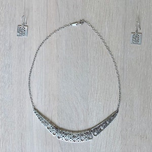 May include: A silver necklace and earrings set with a geometric design featuring a pattern of small circles. The necklace is a curved, crescent shape.