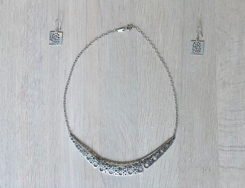 May include: A silver necklace and earrings set with a geometric design featuring a pattern of small circles. The necklace is a curved, crescent shape.