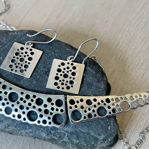 May include: A silver necklace and matching earrings with a geometric design of small circles cut out of the metal. The necklace is curved and the earrings are square.