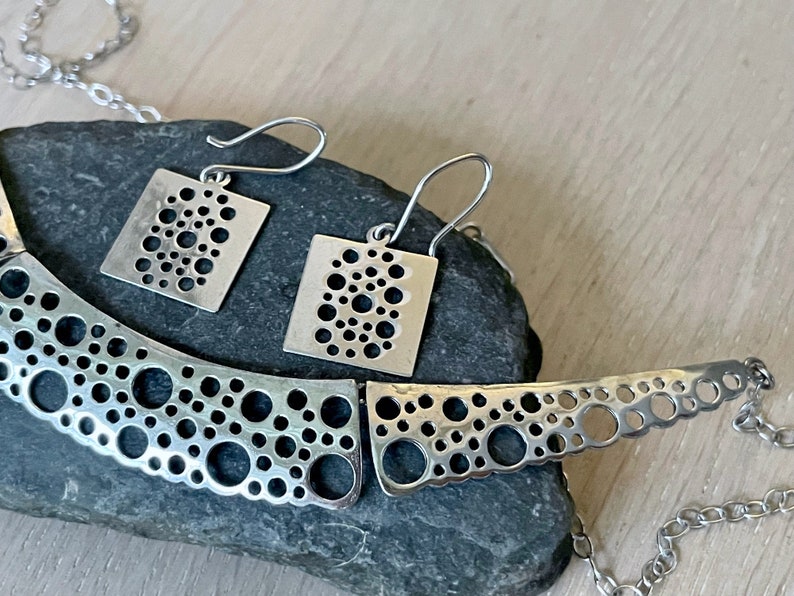 May include: A silver necklace and matching earrings with a geometric design of small circles cut out of the metal. The necklace is curved and the earrings are square.