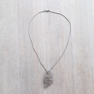 Pode incluir: Um colar prateado com um pendente delicado e vazado. O pendente apresenta um design complexo e interligado, assemelhando-se a uma rede ou a um aglomerado de pequenos ramos. A corrente do colar &eacute; fina e discreta, e o fecho &eacute; vis&iacute;vel no topo.