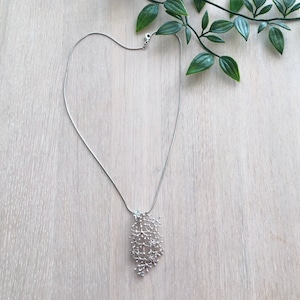 Pode incluir: Um colar prateado com um pendente delicado e ramificado. O pendente apresenta um design org&acirc;nico complexo, que se assemelha a uma rede de pequenos ramos. O colar &eacute; apresentado numa superf&iacute;cie de madeira clara com folhas verdes no fundo.