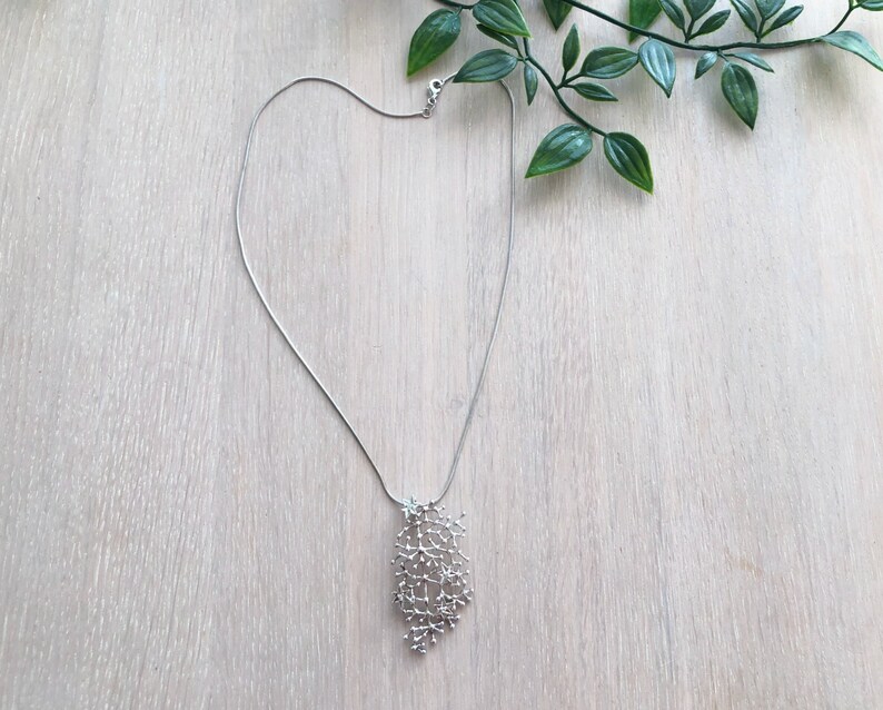 Pode incluir: Um colar prateado com um pendente delicado e ramificado. O pendente apresenta um design org&acirc;nico complexo, que se assemelha a uma rede de pequenos ramos. O colar &eacute; apresentado numa superf&iacute;cie de madeira clara com folhas verdes no fundo.