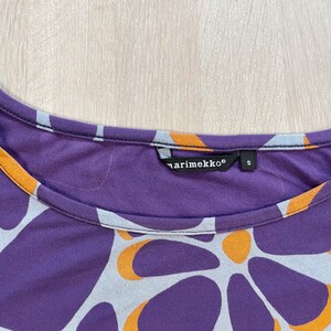 May include: A close-up of a purple, orange, and gray patterned shirt. The shirt has a scoop neck and the brand name "marimekko" is visible on a tag. The pattern consists of large, organic shapes in orange, gray, and purple.