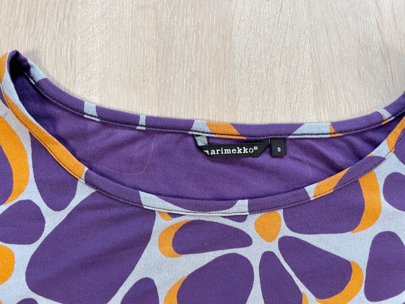 May include: A close-up of a purple, orange, and gray patterned shirt. The shirt has a scoop neck and the brand name "marimekko" is visible on a tag. The pattern consists of large, organic shapes in orange, gray, and purple.