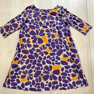May include: A scoop-neck dress with three-quarter sleeves. The dress features a repeating pattern of purple and orange flower shapes on a light gray background. The dress is made of a soft, flowing fabric.
