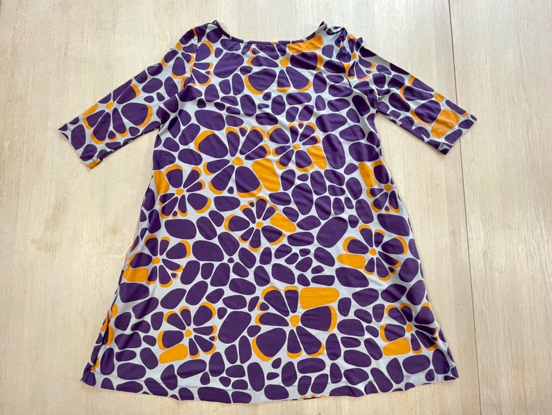 May include: A scoop-neck dress with three-quarter sleeves. The dress features a repeating pattern of purple and orange flower shapes on a light gray background. The dress is made of a soft, flowing fabric.