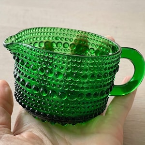 Könnte beinhalten: Ein leuchtend smaragdgrünes Milchkännchen aus Glas mit einer strukturierten Noppenoberfläche auf der Außenseite. Das Gefäß hat einen abgerundeten Körper, einen kleinen Ausguss an einer Seite und einen glatten, geschwungenen Henkel.