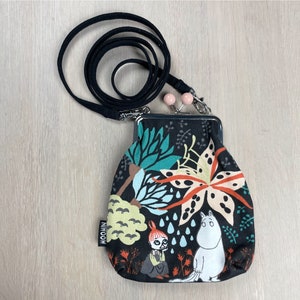May include: A black fabric coin purse with a clasp closure and a black lanyard. The purse features a colorful floral and character design with the word "MOOMIN" on a small tag. The design includes white, orange, and teal elements.