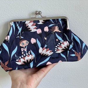 Può includere: Una pochette blu navy con stampa floreale e di personaggi. La pochette presenta una chiusura argentata e un motivo di fiori bianchi, rosa e marroni, con foglie azzurre. I personaggi sono della serie Moomin.