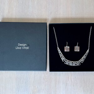 May include: A black box with the text "Design Lilisa Vitali" on the front. The box contains a silver necklace and earrings with a geometric design.