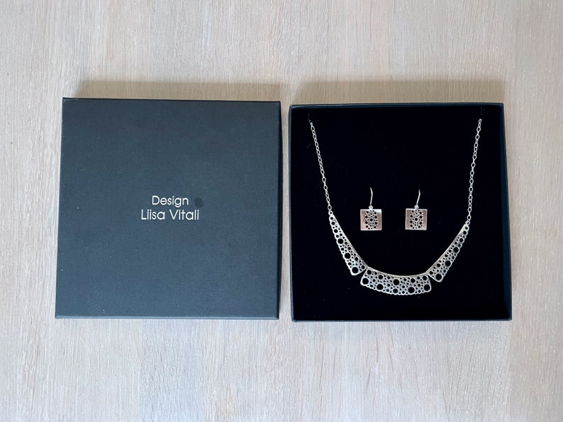 May include: A black box with the text "Design Lilisa Vitali" on the front. The box contains a silver necklace and earrings with a geometric design.