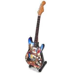 Può includere: Una chitarra elettrica nera con una grafica blu, rossa e bianca di un uomo con un viso di teschio. La chitarra ha un manico in legno ed è su un supporto nero. Il testo "Iron Maiden" è stampato sulla chitarra.