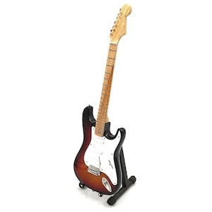 Può includere: Una chitarra elettrica nera e bianca con finitura sunburst su un cavalletto nero. La chitarra ha un manico in acero e un battipenna bianco.