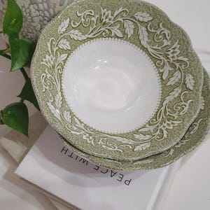 May include: Two white and green patterned ceramic bowls. The bowls have a scalloped edge and a detailed floral design. The bowls are stacked on top of each other. The text "PEACE WITH" is visible on a book beneath the bowls.