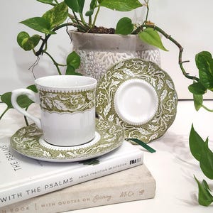 Puede incluir: Un juego de taza y platillo blancos con un diseño floral verde y blanco. La taza está sobre un platillo, con otro platillo cerca. El juego está sobre una pila de libros, con una planta en maceta en el fondo. Los libros tienen el texto "THE PSALMS" y "ABIDE CHRISTIAN MEDITATION".