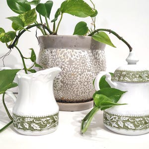 Może przedstawiać: Biały ceramiczny zestaw do śmietanki i cukiernicy z zielonym kwiatowym wzorem. Śmietanka ma uchwyt i falisty brzeg. Cukiernica ma pokrywkę i dwa uchwyty. Roślina doniczkowa z zielonymi liśćmi znajduje się w tle.